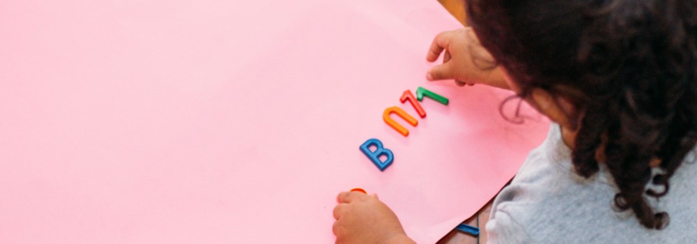 Uma menina está sentada no chão. Ela brinca com letras de plástico em uma cartolina rosa. Imagem ilustra a notícia "Seminário discute a avaliação da qualidade na Educação Infantil".