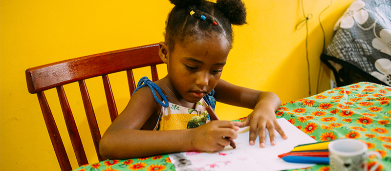 Uma menina negra desenha em uma folha utilizando giz de cera. A imagem ilustra a notícia "Fundação e Alma Preta Jornalismo anunciam série de reportagens sobre a primeira infância negra".