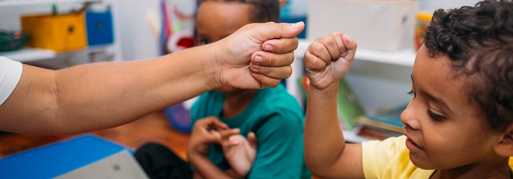 Um menino negro e um profissional da educação infantil sem gênero especificado brincam com as mãos. Imagem ilustra a matéria "Recomendações mostram o que candidatas e candidatos devem fazer para priorizar a primeira infância".