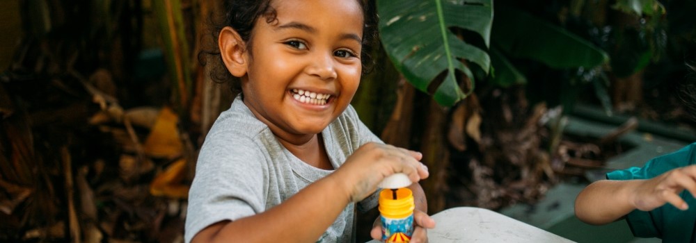 Uma menina negra sorri enquanto segura um frasco de bolhas de sabão. A imagem ilustra notícia que fala sobre diretrizes de qualidade e equidade na educação infantil.