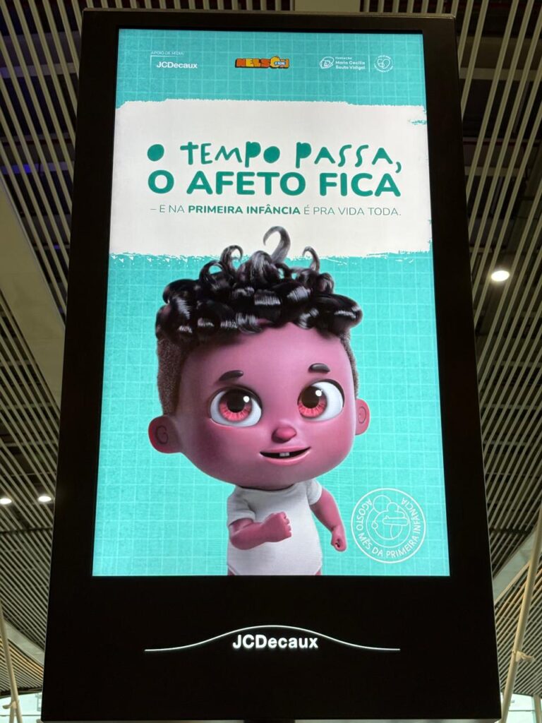 Imagem de Nelson, o Nenê, em uma tela digital. Nelson é um personagem negro de dois anos, com cabelos cacheados e em 3D, que sorri e aponta para o texto "O tempo passa, o afeto fica - e na primeira infância é pra vida toda".