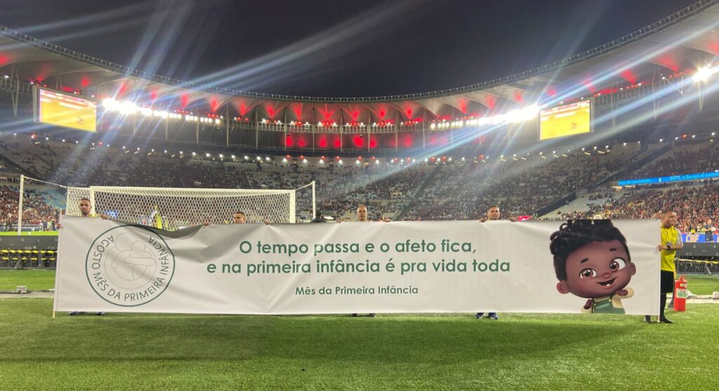 Faixa com Nelson, o nenê, e os dizeres em destaque: "O tempo passa e o afeto fica, e na primeira infância é pra vida toda - Mês da primeira infância". A faixa está estendida pela equipe do time Flamengo, no estádio do maracanã.