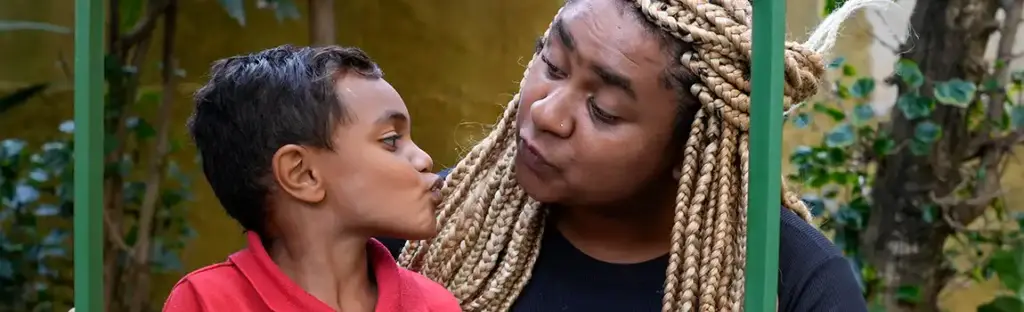 Uma criança pequena com cabelo escuro curto e camiseta vermelha se inclina para beijar sua mãe no rosto, uma mulher negra com longas tranças loiras e uma blusa preta. Eles seguram uma moldura de metal verde que cria uma janela ao seu redor. Ao fundo, temos uma folhagem desfocada, sugerindo um ambiente ao ar livre como um quintal.