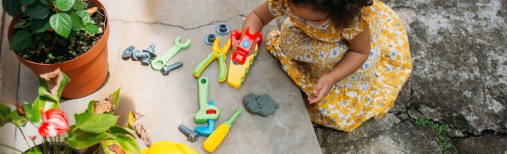 Criança com vestido amarelo floral brincando no chão com brinquedos de ferramentas de plástico coloridos ao lado de um vaso de plantas.