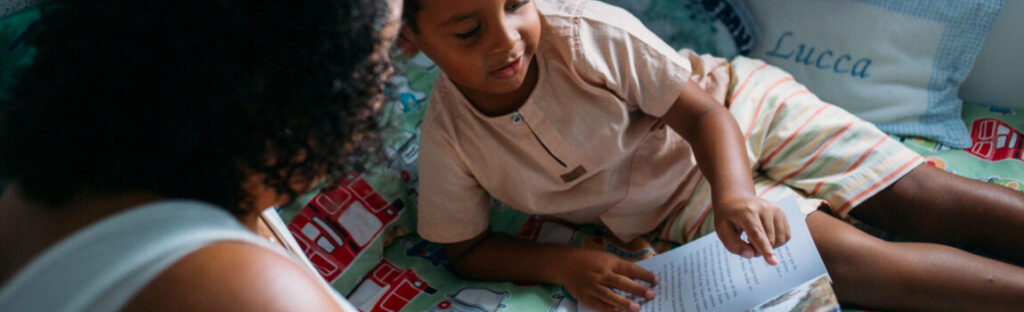 Uma mãe lê para seu filho, que segura um livro deitado na cama. Ilustra matéria sobre campanha de distribuição de kits de leitura realizada pela Fundação Maria Souto Vidigal e Instituto Mauricio de Souza.