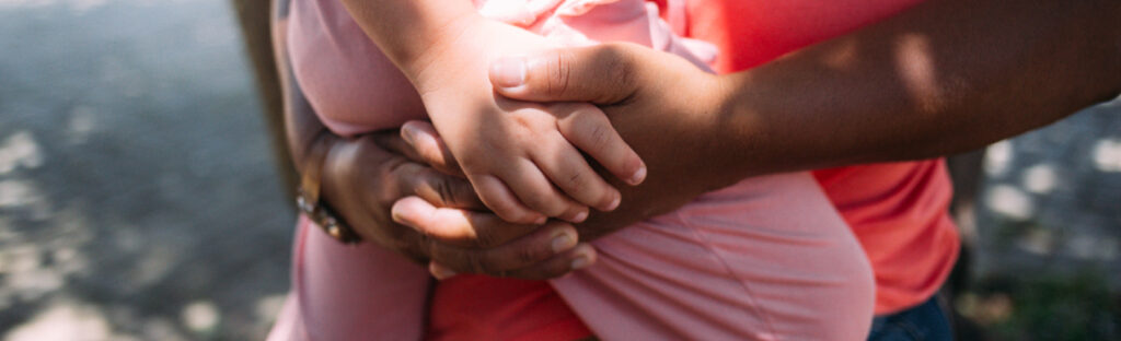 Close das mãos de uma pessoa adulta segurando uma criança no colo, em um gesto de proteção e acolhimento.