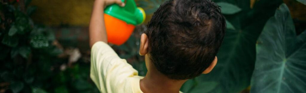 Criança com camiseta amarela rega plantas em um jardim usando um regador colorido.