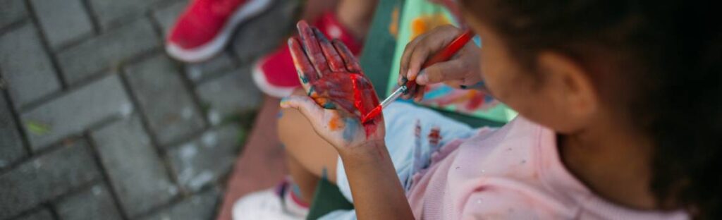 Criança pinta a palma da própria mão com tinta vermelha durante atividade artística ao ar livre.