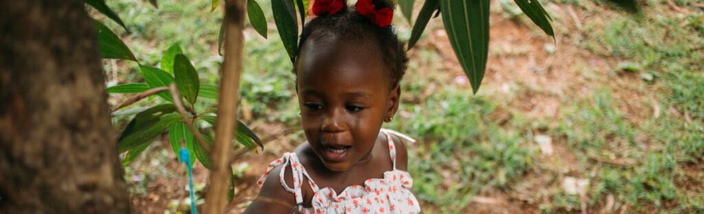 Menina pequena com laços vermelhos no cabelo explora um brinquedo pendurado em um galho, brincando ao ar livre em meio às plantas
