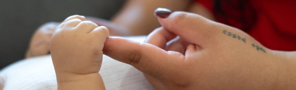 Mão de um bebê segura o dedo de uma pessoa adulta em um gesto de cuidado e vínculo.