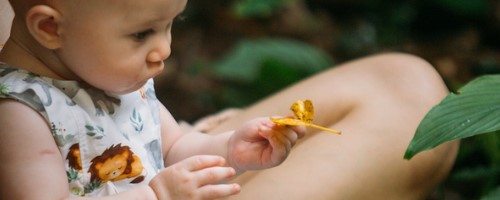 Acompanhado da mãe, um bebê segura uma folha amarela e olha para outra folha verde