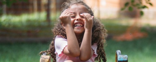 Uma menina de cabelos cacheados sorri. Ela está no topo de um escorregador de madeira.