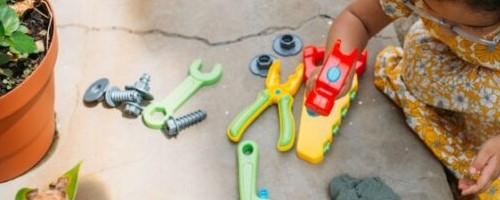 Uma menina com vestido amarelo e florido brinca com ferramentas de plástico próximas a um vaso de plantas