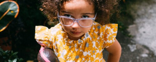 Uma menina parda, de óculos e vestido amarelo, olha com semblante sério para quem a fotografa. Ilustra notícia "Primeira infância e clima - como a crise climática afeta o desenvolvimento infantil"
