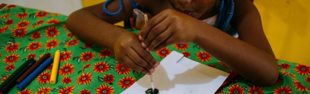 Criança na educação infantil pintando com pincel sobre papel em mesa colorida, com materiais de arte ao redor